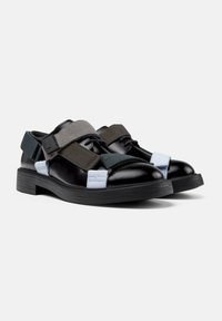 Zapatos de cuero negro con tiras elásticas en gris, verde oscuro y azul claro. Presentan una puntera redondeada y una suela gruesa de goma negra.