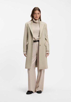 Femme portant un long manteau beige par-dessus un pull à col roulé clair, un pantalon large beige avec une ceinture noire et des chaussures noires, debout sur un fond blanc.
