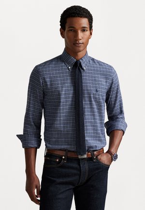 Marineblauwe geruite overhem bij met lichte blauwe accenten, gecombineerd met een donkere spijkerbroek en een bruine riem. Katoenen stof; overhemd heeft een button-down kraag.