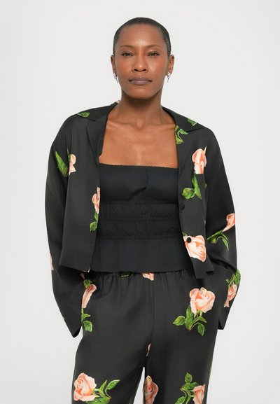 Giacca nera con stampa floreale di rose e foglie verdi, indossata sopra un top senza spalline nero. Tessuto morbido, vestibilità comoda, pantaloni floreali abbinati.