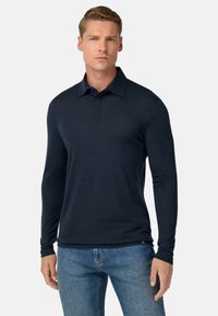 Hombre con una camiseta polo de manga larga, de color azul marino oscuro, y jeans azules, de pie contra un fondo liso y claro.