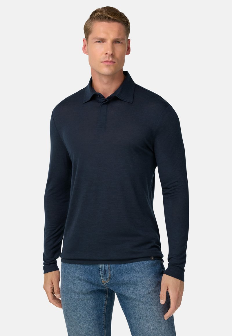 Hombre con una camiseta polo de manga larga, de color azul marino oscuro, y jeans azules, de pie contra un fondo liso y claro.