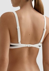 Bela modrček z nastavljivimi naramnicami, črnim trakom in minimalističnim dizajnom. Gladek otip in zapiranje z zaponko na hrbtni strani.