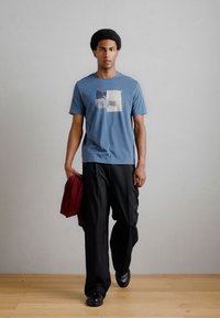 Camiseta de algodão azul com um padrão geométrico, combinada com calças pretas largas e segurando um suéter vermelho-escuro, contra um fundo neutro.