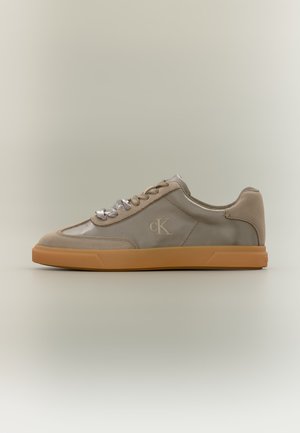 Baskets basses beiges avec semelle en caoutchouc, accents en daim, lacets en satin et logo "cK" sur le côté sur un fond uni.