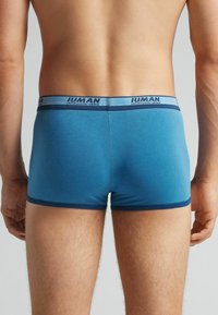Blaue Baumwoll-Boxershorts mit einem kontrastierenden dunkelblauen Bund, der Logo-Text aufweist. Glatte Textur, entworfen für Komfort und Passform.