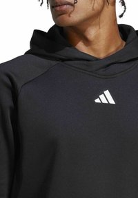 Schwarzer Hoodie mit strukturiertem Finish, ausgestattet mit einer Kängurutasche und einem weißen gestreiften Logo auf der Brust. Weicher Stoff, lässiges Design.