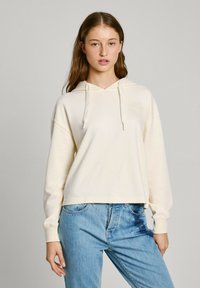 Sudadera crema con cordón, de corte relajado y acentos texturizados. Combinada con jeans de mezclilla azul claro. Diseño simple y contemporáneo.