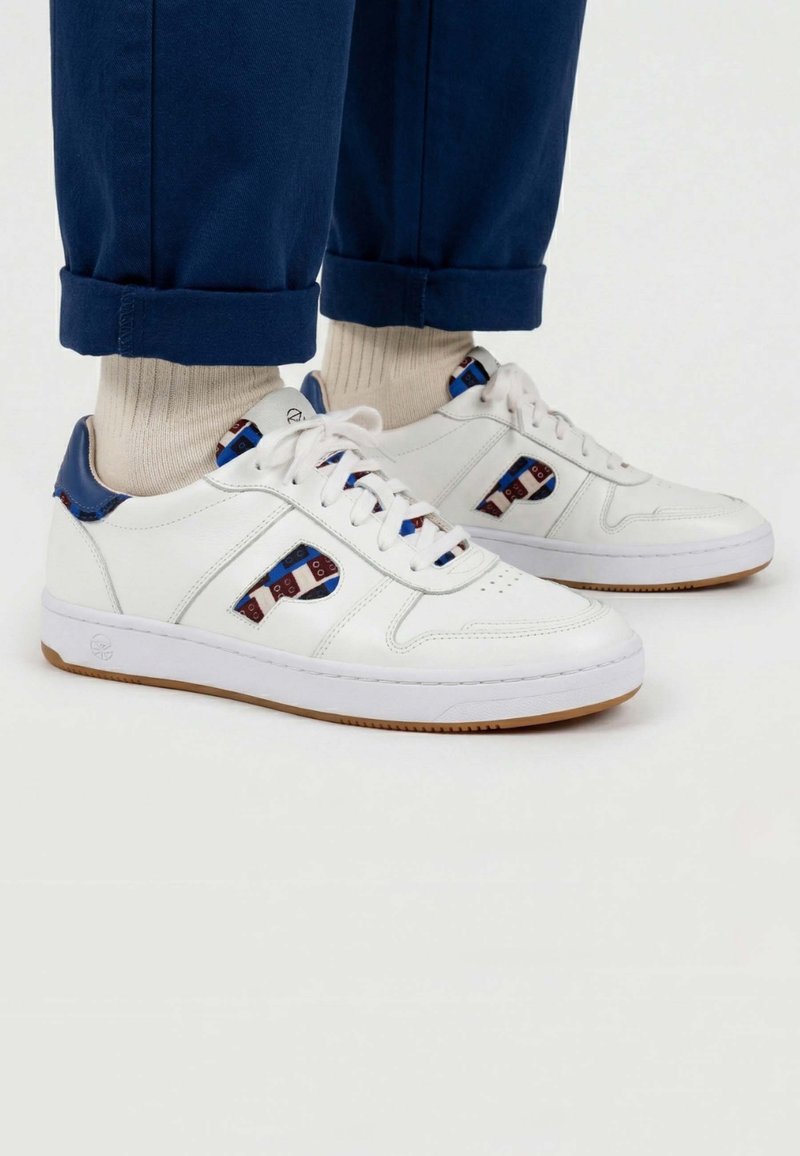 Baskets en cuir blanc avec des détails bleus et rouges à motifs, portées avec des chaussettes beige et un pantalon bleu marine retroussé sur un fond blanc.