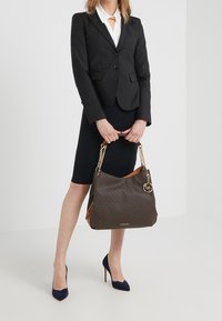 Sac fourre-tout à motif marron avec des poignées en chaîne dorée, présentant une couleur d'accent en haut et une étiquette de marque. Porté avec un costume noir et des talons.