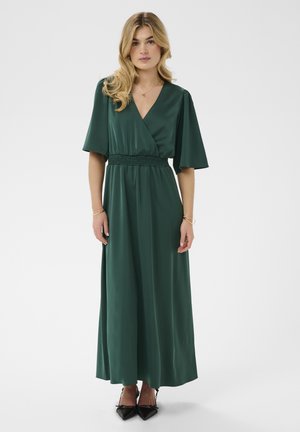 Lokkis blondide juustega naine, kes kannab tumerohelist lühikeste varrukatega V-kaelusega maksikleiti ja musti teravnurkseid kingi, seisab ühevärvilise tausta ees.