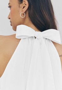 Top bianco con collo halter e un grande fiocco sul retro, realizzato in tessuto morbido e con una linea fluttuante, che mette in risalto una silhouette liscia e pulita.