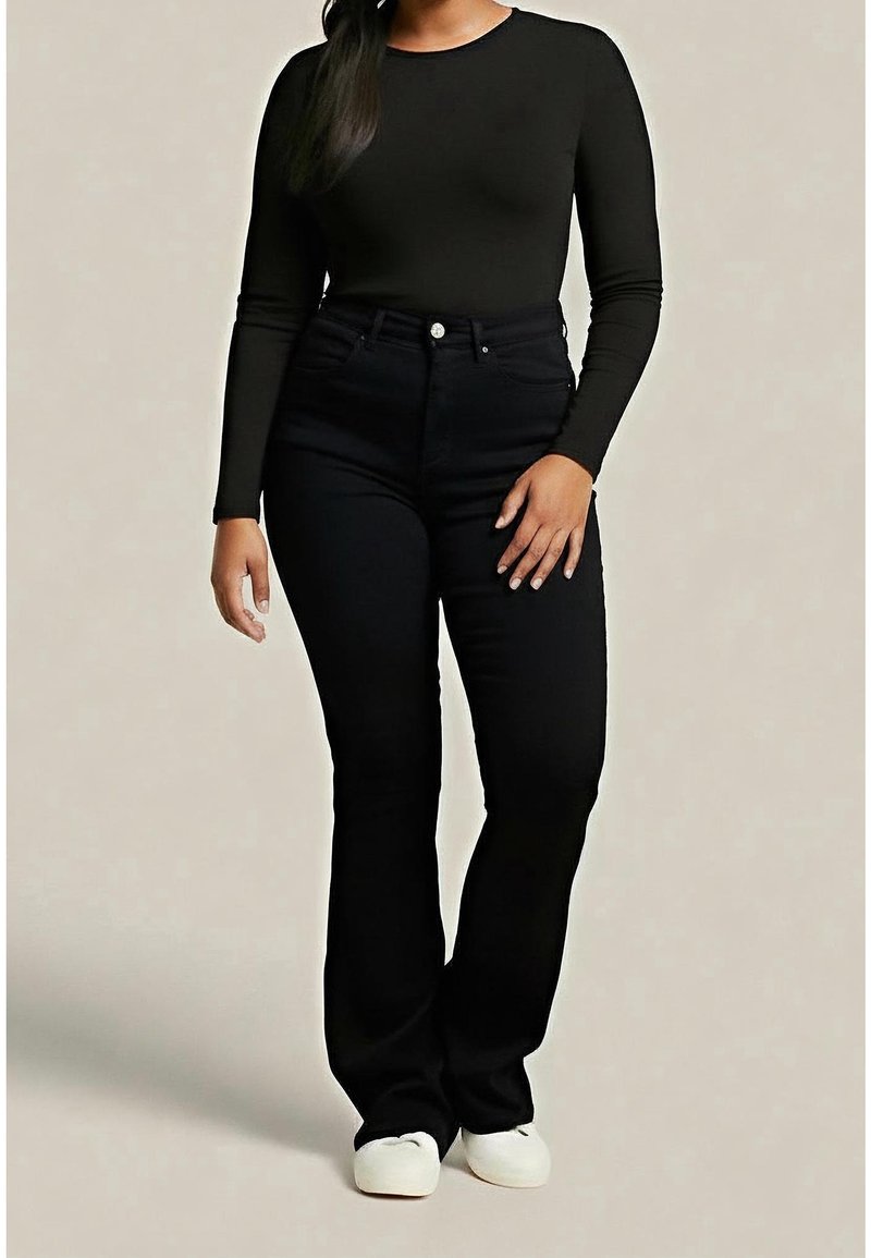 Femme portant un haut noir ajusté à manches longues, un jean noir taille haute et des chaussures blanches, debout devant un fond uni.