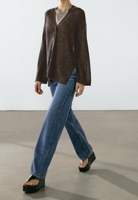 Person wearing a brown buttoned cardigan, blue jeans, black platform sandals, and a layered necklace, walking on a gray floor.