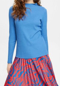 Blå ribbad långärmad topp med en vågig halsringning, kombinerad med en plisserad röd kjol med blå abstrakta mönster.