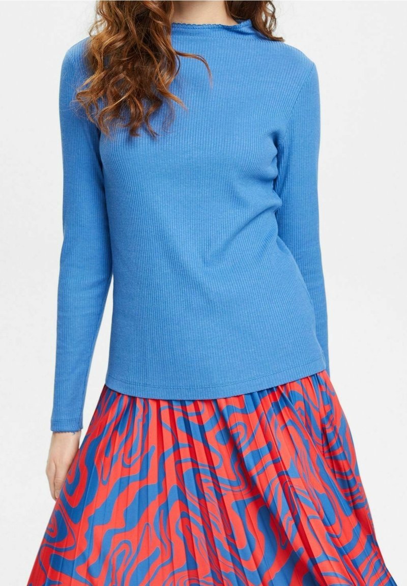 Blå ribbad långärmad topp med en vågig halsringning, kombinerad med en plisserad röd kjol med blå abstrakta mönster.