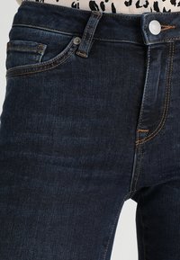 Primer plano de unos jeans de mezclilla de color azul oscuro con costuras naranjas, bolsillos frontales, pasadores de cinturón y un botón plateado en la cintura.