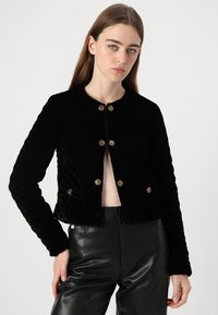 Giacca in velluto trapuntato nera con scollatura rotonda, bottoni decorativi dorati e maniche lunghe, abbinata a pantaloni in pelle nera.