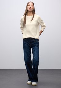 Sudadera crema con puños acanalados, combinada con jeans de pierna ancha de color azul oscuro y zapatillas blancas. El conjunto es sencillo y casual, con un corte relajado.