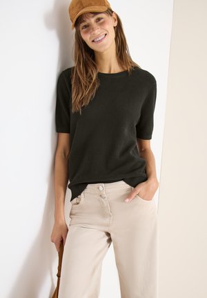 Mujer joven con gorra marrón, camiseta negra de manga corta con textura y pantalones beige, sonriendo con una mano en el bolsillo y apoyándose en una pared blanca.