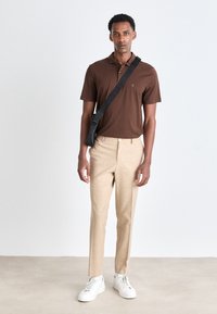 Brun kortermet polo skjorte, beige skræddersyede bukser og hvide sneakers. Sort crossbody taske tilføjer kontrast. Glat stoftekstur.