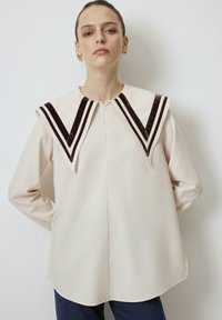 Crème-kleurige blouse met een grote, gestructureerde kraag met donkerbruine fluwelen strepen. Lange mouwen en een sluiting aan de voorkant. Gladde stoftextuur.