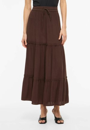 Femme portant une longue jupe marron à volants avec taille à cordon et mules à talons ouvertes noires, debout devant un fond blanc.