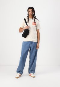 Beige t-skjorte med kort erm og rød grafikk, lyseblå løstsittende jeans og hvite joggesko med røde striper. Svart crossover-veske.