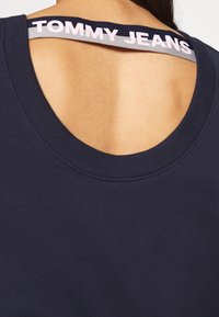 Marineblå sweatshirt med rund halsringning och öppen rygg, med en synlig logoband som säger "TOMMY JEANS" i kontrasterande färger.