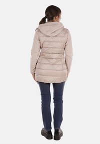 Giacca trapuntata beige chiaro con cappuccio, caratterizzata da cuciture verticali, un taglio lungo e una texture liscia e lucida. Indossata con jeans scuri e stivali robusti.