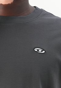 T-shirt en coton noir avec un col rond, arborant un petit logo brodé en blanc et gris sur la partie gauche de la poitrine.