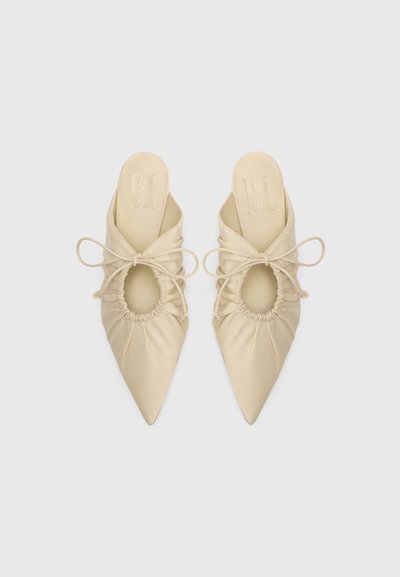Mules de piel beige con punta afilada, detalle fruncido y corte ovalado. Cuenta con cordones finos para ajuste. Textura suave.
