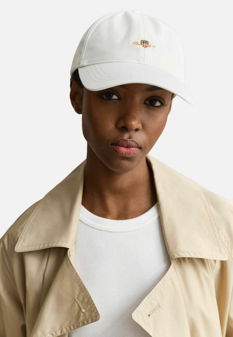 Femme portant une casquette de baseball blanche avec un logo brodé, un trench beige et un t-shirt blanc côtelé, regardant droit devant avec une expression neutre.
