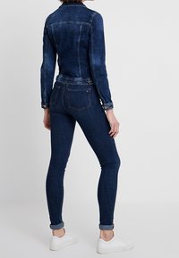 Tmavě modrá džínová bunda s přiléhavým střihem, zapínáním na knoflíky a kapsami. Kombinována s tmavými úzkými džínami a bílými teniskami.