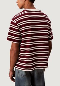 Proužkované tričko v barvě vína a bílé, krátké rukávy, kulatý výstřih, volný střih, bavlněný materiál, viditelné švy, nošeno s lehkými džíny.