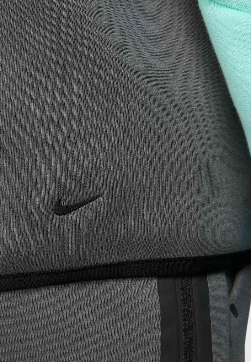 Top atlético gris con el logo negro de Nike. Presenta acentos verdes, textura suave y un diseño ajustado. Incluye una banda negra en el dobladillo.