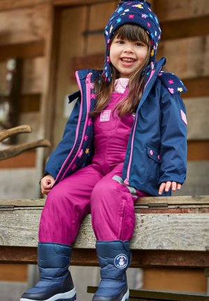 Kind in een blauw en roze gestreepte winterjas, een roze sneeuwpak en blauwe laarzen met witte zolen, zittend op een houten bank.