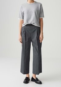Personne portant une chemise grise à manches courtes, un pantalon gris à larges jambes taille haute à rayures fines et des mocassins noirs sur un fond uni.