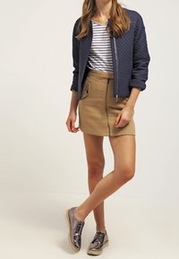 Veste bomber matelassée bleu marine, chemise rayée blanche et bleu marine, jupe beige zippée et chaussures lacées métalliques grises avec une finition texturée.