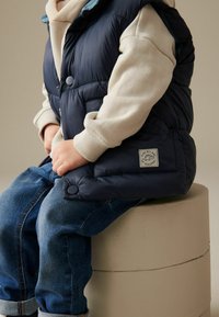 Gilet imbottito blu navy con chiusure a bottone, realizzato in tessuto testurizzato, indossato sopra una felpa beige chiaro e jeans blu, seduto su uno sgabello cilindrico.