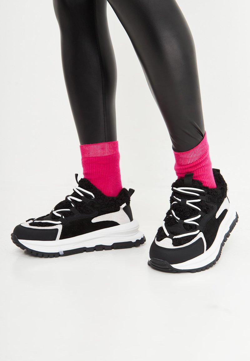 Sneakers in bianco e nero con accenti in peluche, calzini a coste rosa acceso e una suola robusta e texturizzata. Il design enfatizza comfort e stile.