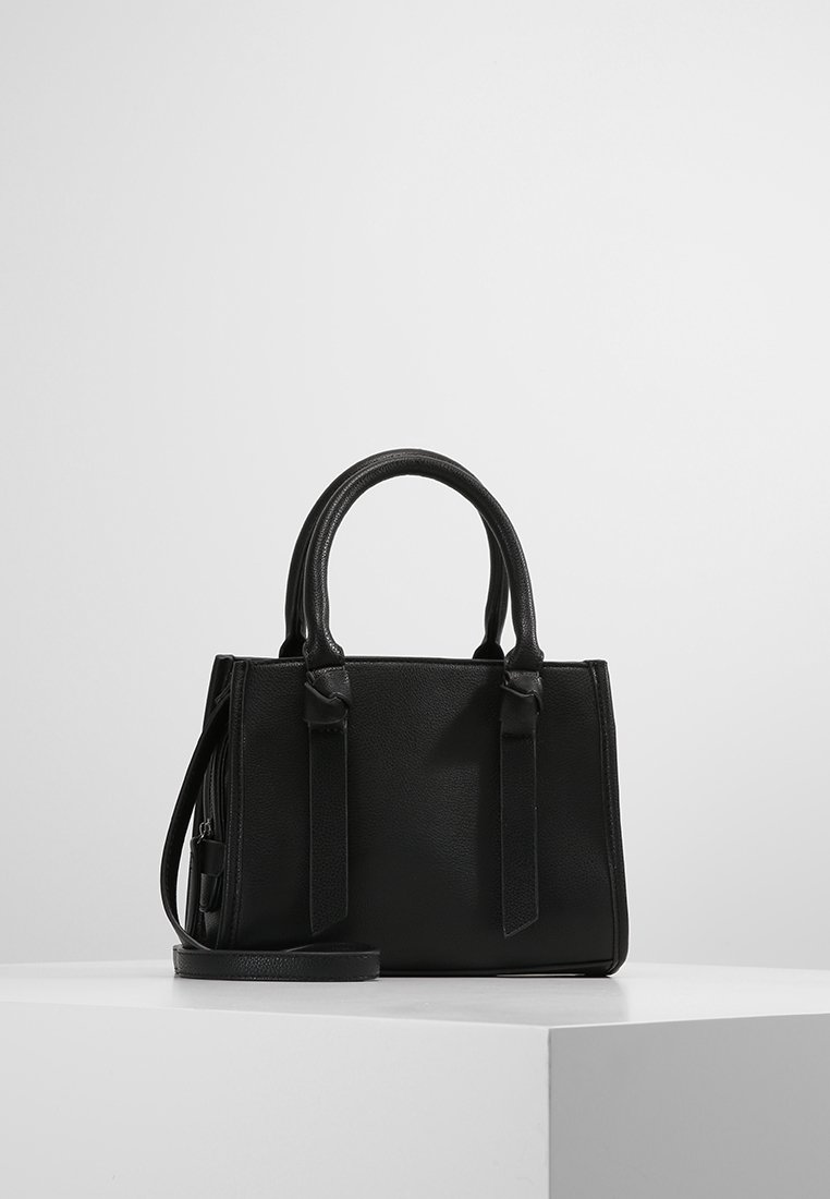 Sac à main en cuir noir avec doubles poignées et bandoulière amovible, posé sur une surface blanche sur un fond uni clair.