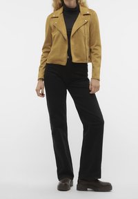 Suède motorjack in mosterdgeel, met een ritssluiting, zijzakken en een getailleerd ontwerp, gecombineerd met zwarte uitlopende broeken en donkere laarzen.