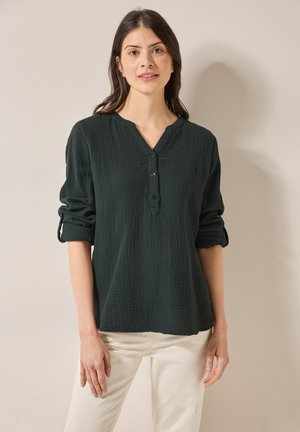 Mörkgrön, texturerad blus med lång ärm, V-ringning och knapplist. Uppvikta ärmar med knappdetalj. Matcher med ljusa byxor.