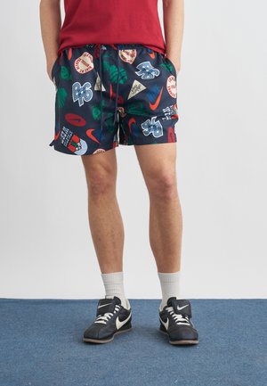 Hombre con pantalones cortos azul marino con logotipos gráficos coloridos, camiseta roja, calcetines blancos y zapatillas negras Nike de pie sobre suelo azul.