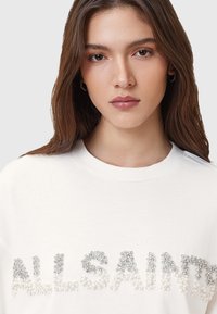 Biele bavlnené tričko s nápisom "ALLSAINTS" vytvoreným z rôznych veľkostí kamienkov a korálok, ktoré tvoria textúrovaný, zdobený dizajn.