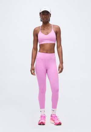 Mujer con sujetador deportivo rosa, leggings a juego, gorra blanca de Nike, calcetines blancos y zapatillas de running rosas, de pie frente a un fondo blanco.