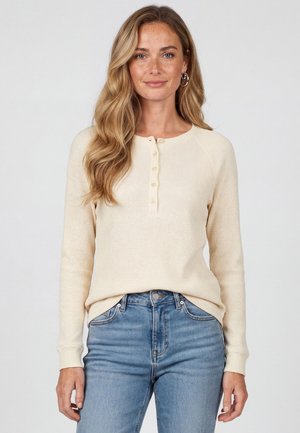 Kvinde med langt bølget blondt hår iført øreringe, en cremefarvet knaplukket langærmet sweater og blå jeans, stående mod en ensfarvet baggrund.