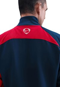 Die Nike-Jacke hat einen roten oberen Bereich und einen marineblauen Körper. Sie verfügt über ein weißes Logo auf der Rückseite und hat eine glatte, atmungsaktive Textur.