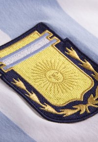 El escudo de armas bordado presenta un sol amarillo en el centro, rodeado de acentos azules y dorados sobre un fondo a rayas de color azul claro.